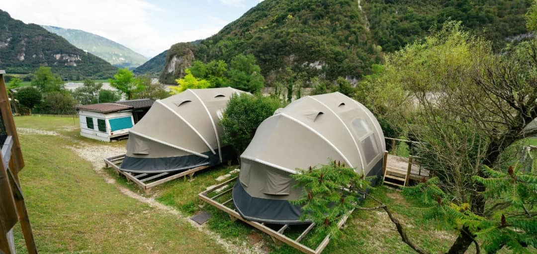Zwei Big Oak Lodges XS mit kuppelförmiger Zeltplane auf Holzterrassen, inmitten grüner Berge mit Felswänden