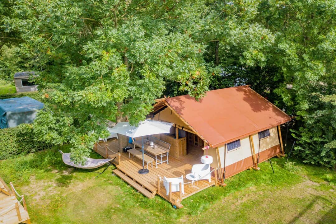 Luftaufnahme der Cozy Lodge mit rotbraunem Dach unter hohen Bäumen, mit Sonnenschirm, Esstisch und Hängematte auf der Veranda