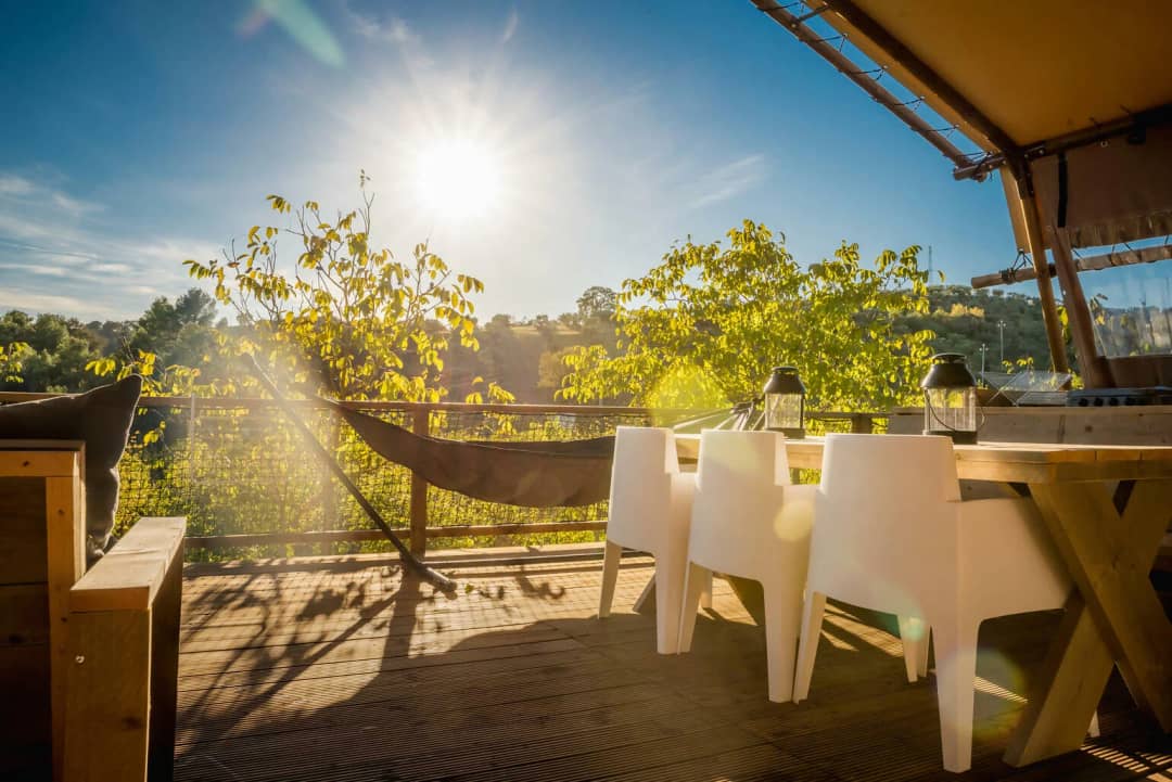 Ein Esstisch aus Holz mit gekreuzten Beinen und weißen Stühlen auf der Terrasse der Desert Lodge XL, während die Sonne über den Bäumen steht