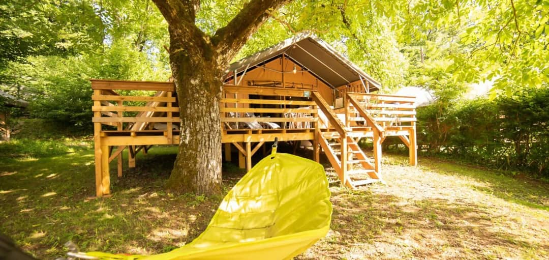 Eine Safari-Lodge auf Stelzen unter einem großen Baum, mit einer Holztreppe zur Terrasse und einer gelben Hängematte im Vordergrund