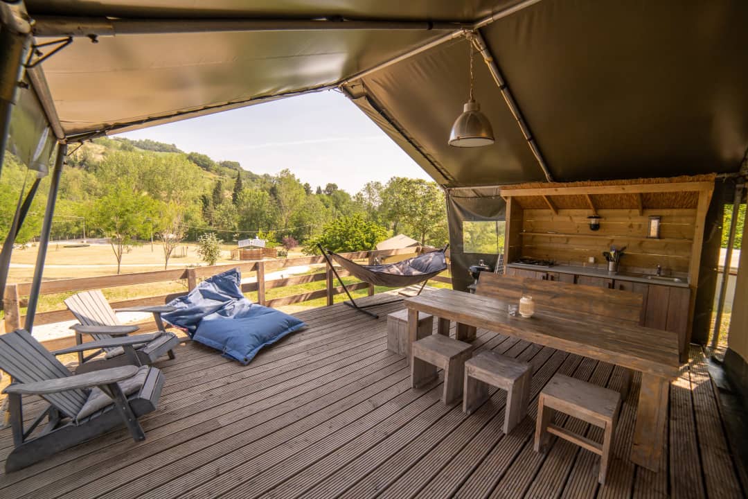 Veranda der Safari Lodge für 2 bis 6 Personen mit Gartenstühlen, blauen Sitzsäcken, einer Hängematte und einem langen Tisch mit Hockern vor einer Rasenfläche